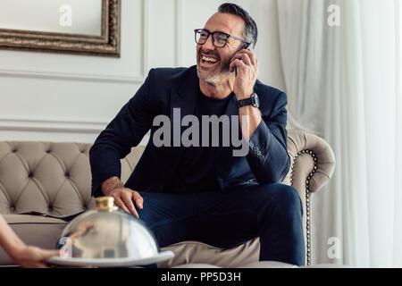 Allegro imprenditore parlando al telefono cellulare in camera in hotel con un cameriere che serve cibo. Servizio in camera Servizio cameriere che serve cibo a un imprenditore di ridere su h Foto Stock