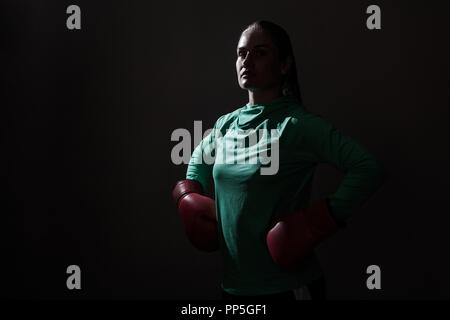 Silhouette Ritratto di giovane donna boxer in green sport usura e Guanti rossi in piedi e guardando la fotocamera con le mani sulla cintura e orgoglioso volto coperto Foto Stock