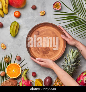 Le mani di una donna tenendo un marrone piastra vuota attorno a una diversa frutta tropicali mature su un cemento grigio Sfondo con copia spazio. Lay piatto Foto Stock