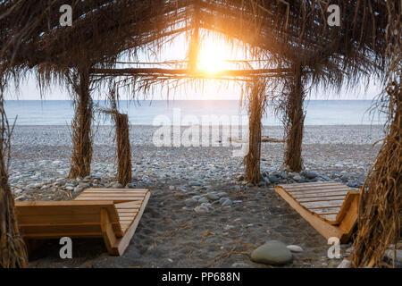 Coppia di lettini di legno sotto una tettoia di canna di tetto sulla spiaggia al tramonto Foto Stock