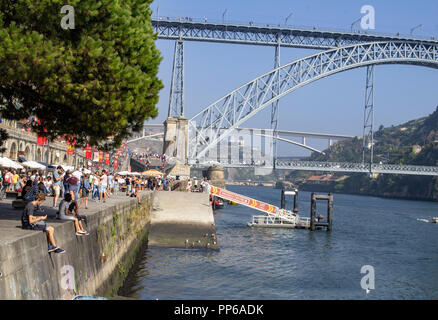 Porto, Portuga. I turisti costeggiano le banchine lungo il fiume Duoro, con il Ponte Maria Pia sullo sfondo e un piccolo motoscafo che naviga sul fiume. Foto Stock