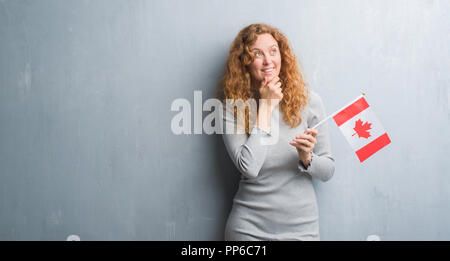 Giovane donna rosso su grigio parete grunge holding bandiera del Canada faccia seriamente pensando di domanda, molto confusa idea Foto Stock