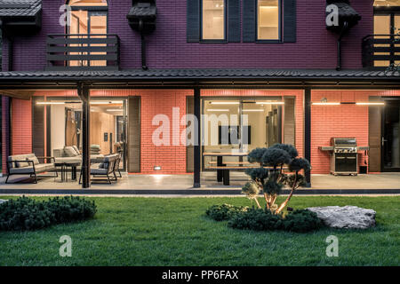 Moderno edificio di due piani country house con spie luminose. Presenta grandi finestre con porte di vetro, tenda, terrazza con tavoli di legno e panche e Foto Stock