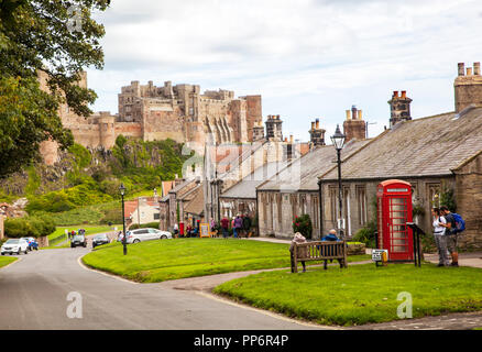 Turisti e villeggianti che lungo la strada principale del villaggio di Northumberland Bamburgh England Regno Unito con il castello di Bamburgh in background Foto Stock