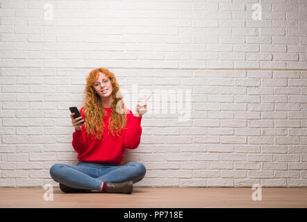Giovani redhead donna seduta su un muro di mattoni a parlare al telefono molto felice rivolto con la mano e le dita per lato Foto Stock
