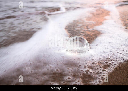 Schiuma e Wave Wrap Around Palla di vetro seduto sulla sabbia nella luce del mattino Foto Stock