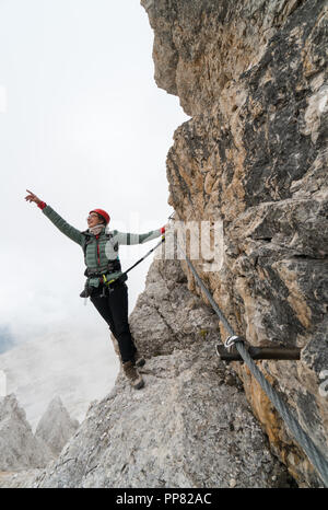 Giovane femmina attraente studentessa universitaria su un piano verticale ed esposta di roccia si arrampica su una via ferrata mentre sorridente e puntamento Foto Stock