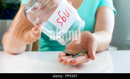 Primo piano della giovane donna versando poche monete a portata di mano dal vaso di vetro con qualche risparmio di denaro Foto Stock