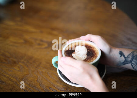 Una femmina con un braccio tatuaggio siede a un tavolo di legno tenendo una tazza di cappuccino in una tazza verde Foto Stock