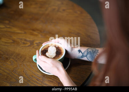 Una femmina con un braccio tatuaggio siede a un tavolo di legno tenendo una tazza di cappuccino in una tazza verde Foto Stock