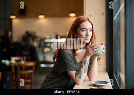 Testa rossa ragazza con braccio tatuaggio, seduto da solo in una caffetteria guardando attraverso la finestra, Liverpool, Regno Unito Foto Stock