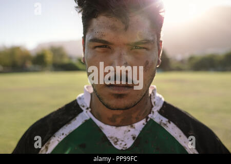 Close up ritratto del giocatore di rugby con fango viso e uniforme sul campo. Giovane atleta a fissare la fotocamera sul terreno. Foto Stock