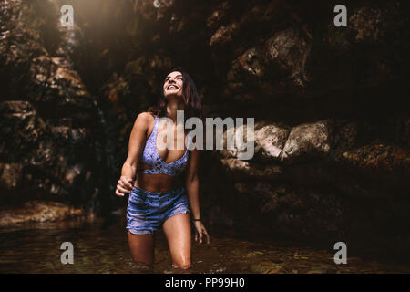 Giovani caucasici donna bella turista in una cascata di stagno nella giungla cercando e sorridente. Bella ragazza che indossa un bikini top e pantaloncini in stagno Foto Stock