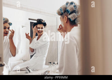 Due donne in accappatoi di applicare la crema sulla faccia in piedi di fronte a specchio. Le donne con rulli di arricciatura appuntato sui capelli toelettatura a casa. Foto Stock