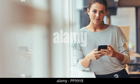 Imprenditrice in piedi da una finestra controllo il suo telefono cellulare. Donna prendendo pausa dal lavoro in piedi con il cellulare in mano. Foto Stock