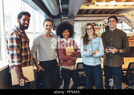 Felice i colleghi in piedi in office holding laptop e tablet. Gruppo multietnico di colleghi di lavoro in piedi insieme in ufficio. Foto Stock