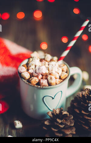Il Natale di cioccolata calda con marshmallows su una tavola in legno rustico con pigne nelle quali, rosso calze di lana e le luci rosse bokeh di fondo. Messa a fuoco selettiva Foto Stock