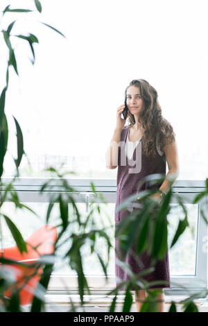 Ritratto di giovane contemporanea donna che indossa abiti comodi che parla al telefono e guardando la telecamera mentre in piedi in camera con le piante Foto Stock