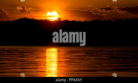 Arancione tramonto attraverso le nuvole, oltre la sponda opposta riflettendo sull'acqua. Foto Stock