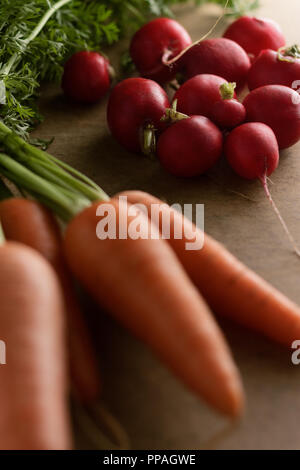 Close-up di un mazzetto di ravanelli, circondato da carote fresche e una testa di lattuga su una luce marrone a bordo, sotto la luce del mattino. Foto Stock