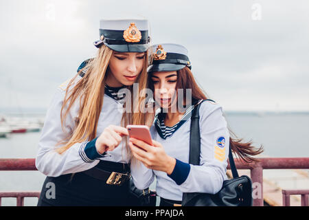 Due college studenti donne di Marine Academy utilizza lo smartphone per mare indossando uniforme. Amici messaggio di lettura da coste Foto Stock