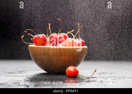 Foto di ciliegia di colore rosso nella tazza in legno sulla tabella grigia, sfondo nero Foto Stock