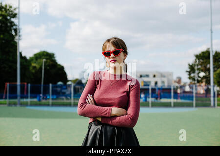 Moda sparare con una ragazza che indossa un marrone rossiccio superiore e una gonna nera in un parco di pallacanestro Foto Stock