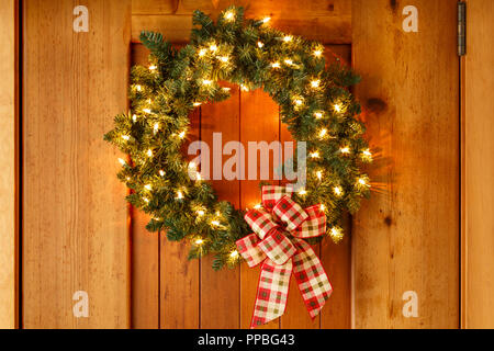 Bellissime decorazioni di Natale corona con nastro di prua e luci su legno porta anteriore dello sfondo. Semplice e rustico stile country holiday home decor. Foto Stock