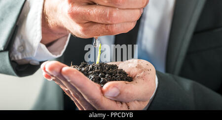 Imprenditore tenendo una pianticella di germogliazione in terra fertile cupped nel palmo della sua mano irrigazione con gli altri in una immagine concettuale di environme Foto Stock