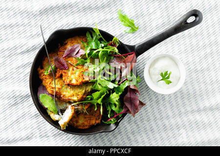 Comfort food concept. Zucchine fritte frittelle servite in piccoli ghisa padella con insalata su sfondo bianco con yogurt greco o panna acida top Foto Stock