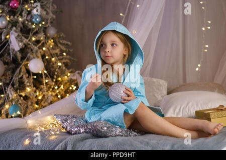 Bambina in accappatoio si siede sul letto e sorrisi. Ella è in possesso di una sfera di vetro nella sua mano. Albero di natale con ornamenti in background. Felice Foto Stock