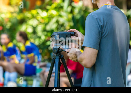 Registrazione Uomo partita di calcio con la sua videocamera su un treppiede in un campo esterno con giocatori sfocata e alberi sullo sfondo Foto Stock