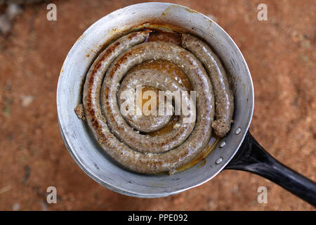 Salsiccia namibiano essendo fritto in un rustico padella su una stufa sul pavimento sabbioso Foto Stock