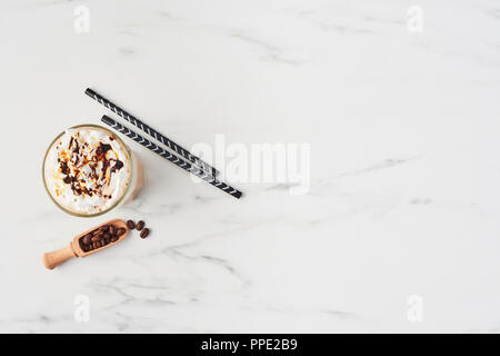 Vista superiore del latte ghiacciata caffè in un bicchiere alto con caramello e sciroppo di cioccolato sulla panna montata. In marmo bianco con sfondo spazio copia. Foto Stock
