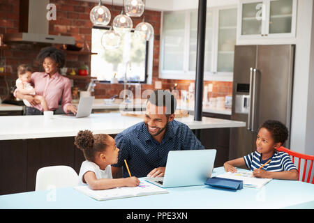 Padre aiuta i bambini con i compiti mentre la madre con bambino utilizza laptop in cucina Foto Stock