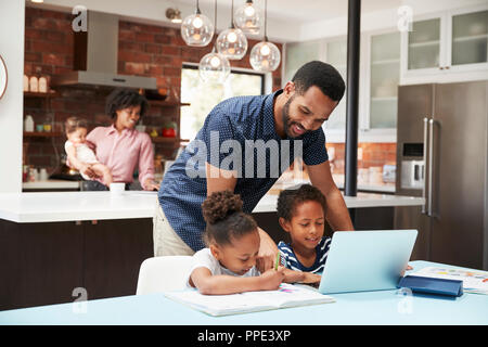 Padre aiuta i bambini con i compiti mentre la madre con bambino utilizza laptop in cucina Foto Stock