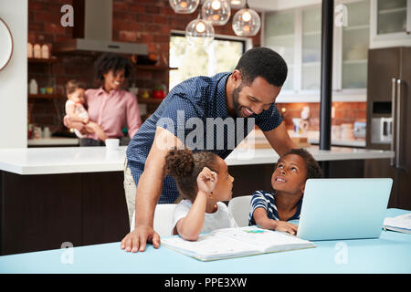 Padre aiuta i bambini con i compiti mentre la madre con bambino utilizza laptop in cucina Foto Stock