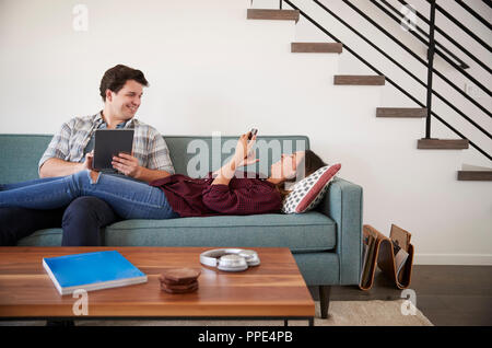 Giovane rilassante sul divano di casa utilizzando il telefono cellulare e la tavoletta digitale Foto Stock