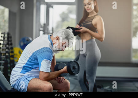 Giovane femmina calda personal trainer senior supporto uomo grigio per la corretta esecuzione di esercizi per mantenere il suo corpo in tono. Uomo anziano fare esercizio con il manubrio, mentre pullman in piedi vicino alla palestra. Foto Stock