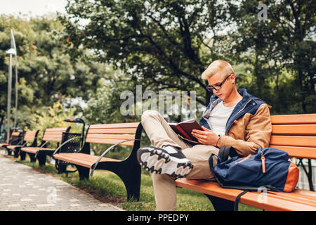 Studente di college con zaino libro lettura passeggiate nel parco di autunno seduta sul banco di lavoro. L'uomo studiare all'aperto Foto Stock