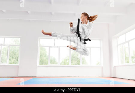 Sportivo da donna in kimono bianco con cintura nera di salto e di eseguire kick. Ragazza giovane con fiducia facendo complessi elementi di tecnica di karate con grave l'espressione del viso. Uno stile di vita sano concetto. Foto Stock