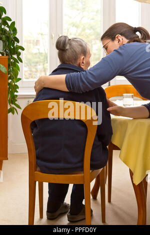 Vista posteriore di un senior donna seduta a casa beeing abbracciato da suo nipote che sta aiutando il suo durante il giorno, eldercare da membri della famiglia Foto Stock