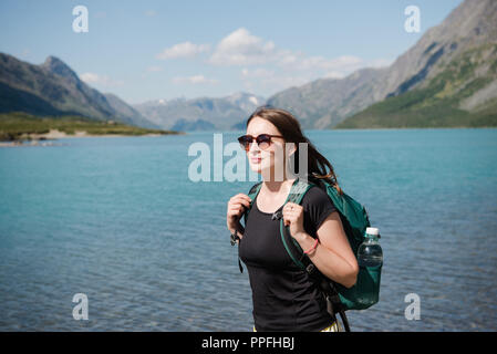 Bella giovane donna in occhiali da sole con zaino in piedi vicino al maestoso Lago Gjende, Besseggen ridge, parco nazionale di Jotunheimen, Norvegia Foto Stock