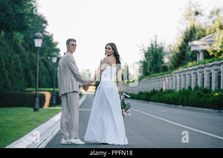 Vista posteriore della sposa e lo sposo Holding Hands, camminando sulla strada e guardando la fotocamera Foto Stock