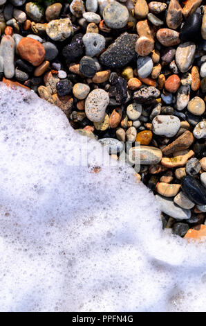 I ciottoli sulla spiaggia coperto per metà dal bianco schiuma di onda Foto Stock