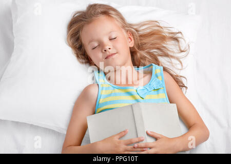 Vista della bella ragazza con lunghi capelli di luce posti letto dopo la lettura di fiaba, mantiene il libro su stomaco, risiede nel comodo letto bianco, è piacevole sogni, e Foto Stock