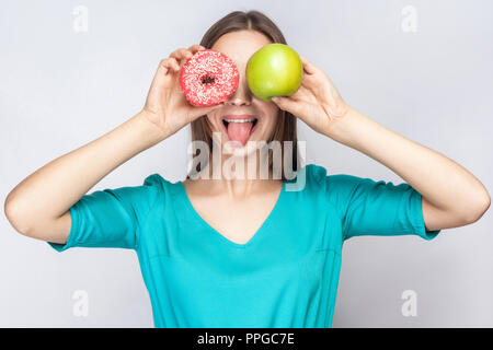 Ritratto di giovane positivo crazy ragazza in blu camicetta in piedi, azienda, copertura rendendo gli occhi con occhiali rosa donut e mela verde e mostra tongu Foto Stock