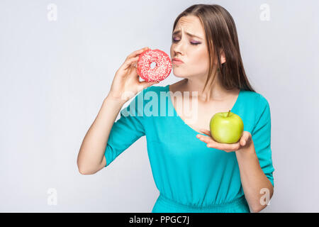 Ritratto di desiderose giovane bella ragazza in blu camicetta in piedi, azienda ciambella rosa e verde mela nelle mani e voleva che scegli unheathy cibo su Foto Stock