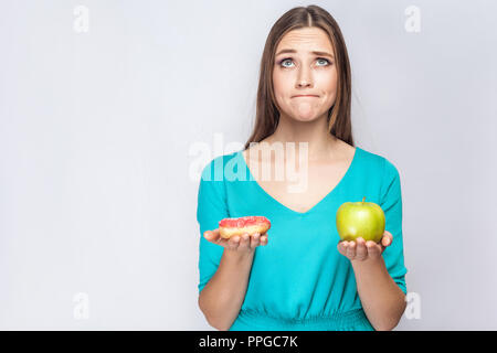 Ritratto di sognare giovane bella ragazza in blu camicetta in piedi, azienda ciambella rosa e verde mela nelle mani come pesatura con faccia intelligentemente o Foto Stock