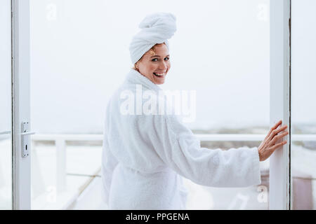 Donna sorridente in un accappatoio con un asciugamano avvolto sulla testa. Vista posteriore di una donna in piedi in balcone dopo il suo bagno. Foto Stock